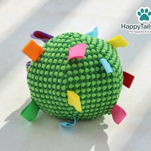 May include: A green, textured ball-shaped pet toy with colorful fabric tags. The toy has a round shape and is covered in small, raised bumps. The tags are in various colors, including orange, pink, yellow, and blue. The HappyTailsShop logo is in the background.