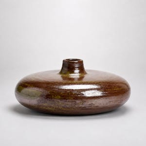 May include: A low-profile, disc-shaped ceramic vase with a glossy, dark brown glaze featuring subtle olive green and bronze undertones. The vessel has a wide, flattened body and a short, narrow cylindrical neck with a small circular opening.