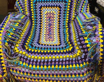 May include: A handmade crochet blanket in a granny square pattern. The blanket features a vibrant color palette of purple, blue, yellow, and green. The edges are finished with fringe in coordinating colors. The blanket is draped over a chair.