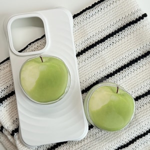 May include: White phone case with a green apple design and a matching phone grip. The case has a swirl pattern. The apple design is a clear, round grip with a green apple inside. The items are on a white and black striped fabric.