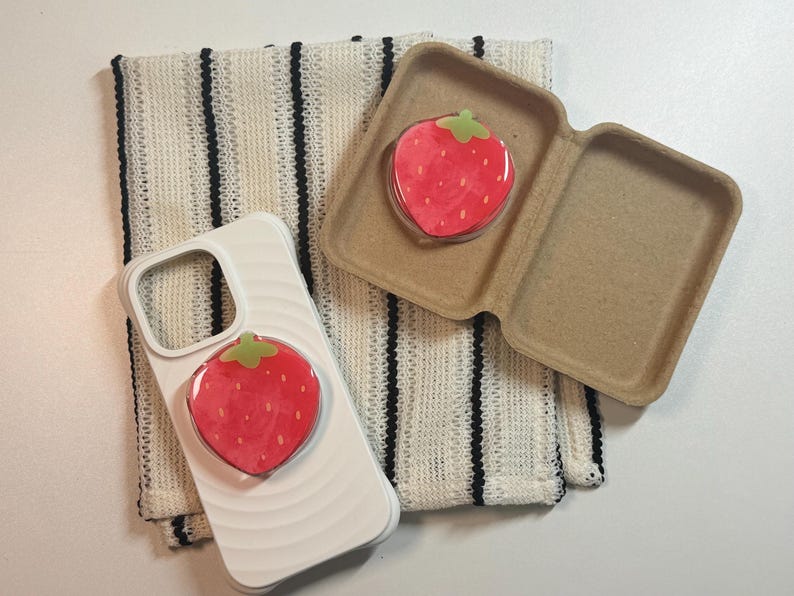 Pu&ograve; includere: Una custodia per telefono bianca con un popsocket a fragola rossa e un popsocket abbinato in una custodia marrone. Un tessuto a righe color crema &egrave; sullo sfondo.