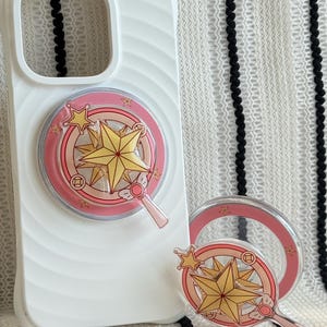 Puede incluir: Funda de teléfono blanca con un diseño de estrella rosa y dorada. El diseño incluye una estrella, círculos y una pequeña varita. Un pop socket a juego está al lado de la funda. El fondo es un patrón de rayas.