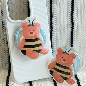 May include: A white phone case with a light blue pop-up grip featuring a cartoon bear dressed as a bee. The bear is peach-colored with black and yellow stripes. A second pop-up grip is also visible.