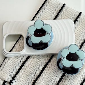Könnte beinhalten: Weißes Handycase mit einem hellblauen, blütenförmigen Katzendesign. Die Katze ist schwarz mit einer hellblauen Blume um ihren Kopf. Das Case und das Katzendesign befinden sich auf einer weiß-schwarz gestreiften Oberfläche.