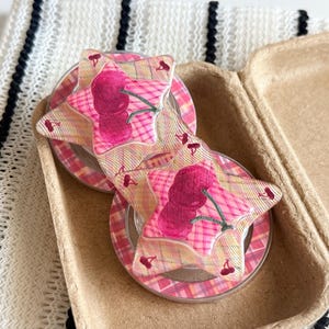 May include: Two star-shaped hair clips featuring a pink and yellow plaid pattern with a dark pink cherry graphic. Each clip has a glossy finish and is layered over a circular base with a matching pink plaid design, creating a layered effect.