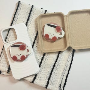 Może przedstawiać: Białe etui na telefon z motywem filiżanki kawy i pasującym uchwytem na telefon. Filiżanka kawy ma uśmiechniętą twarz i czerwone akcenty. Uchwyt na telefon jest przymocowany do etui. Na zdjęciu znajduje się r&oacute;wnież pasujący uchwyt na telefon z filiżanką kawy w kartonowym pudełku.