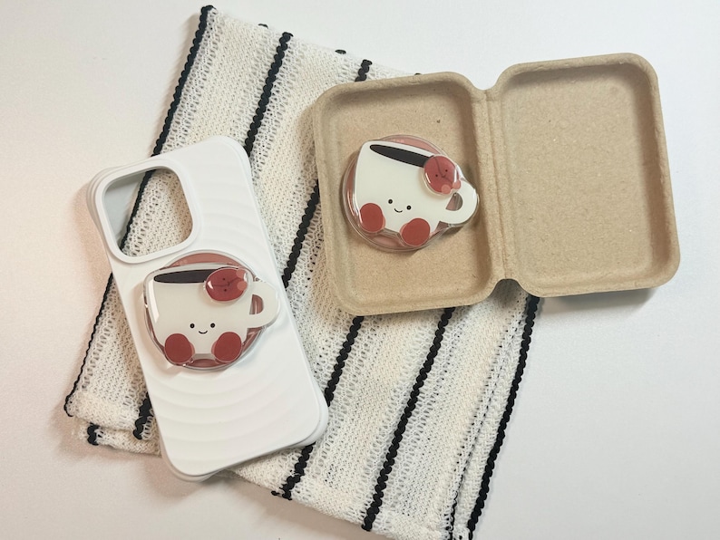 Może przedstawiać: Białe etui na telefon z motywem filiżanki kawy i pasującym uchwytem na telefon. Filiżanka kawy ma uśmiechniętą twarz i czerwone akcenty. Uchwyt na telefon jest przymocowany do etui. Na zdjęciu znajduje się r&oacute;wnież pasujący uchwyt na telefon z filiżanką kawy w kartonowym pudełku.
