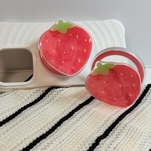 Pu&ograve; includere: Una custodia per telefono bianca con un'impugnatura per telefono a forma di fragola e un pop socket abbinato. I disegni delle fragole sono rossi con cime verdi e piccoli semi gialli. Gli articoli sono su un tessuto a righe bianche e nere.
