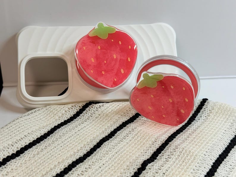 Pu&ograve; includere: Una custodia per telefono bianca con un'impugnatura per telefono a forma di fragola e un pop socket abbinato. I disegni delle fragole sono rossi con cime verdi e piccoli semi gialli. Gli articoli sono su un tessuto a righe bianche e nere.