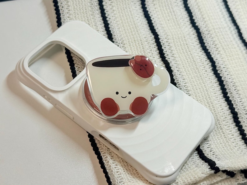 Może przedstawiać: Białe etui na telefon z uroczym uchwytem na telefon w kształcie filiżanki kawy. Uchwyt przedstawia uśmiechniętą filiżankę kawy z czerwonymi akcentami i ziarnem kawy na g&oacute;rze. Etui na telefon leży na biało-czarnej tkaninie w paski.