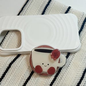 Może przedstawiać: Białe etui na telefon z okrągłym wzorem i uchwytem na telefon w kształcie filiżanki kawy. Uchwyt na telefon jest biały z uśmiechniętą twarzą, czerwonymi akcentami i wzorem ziarna kawy.