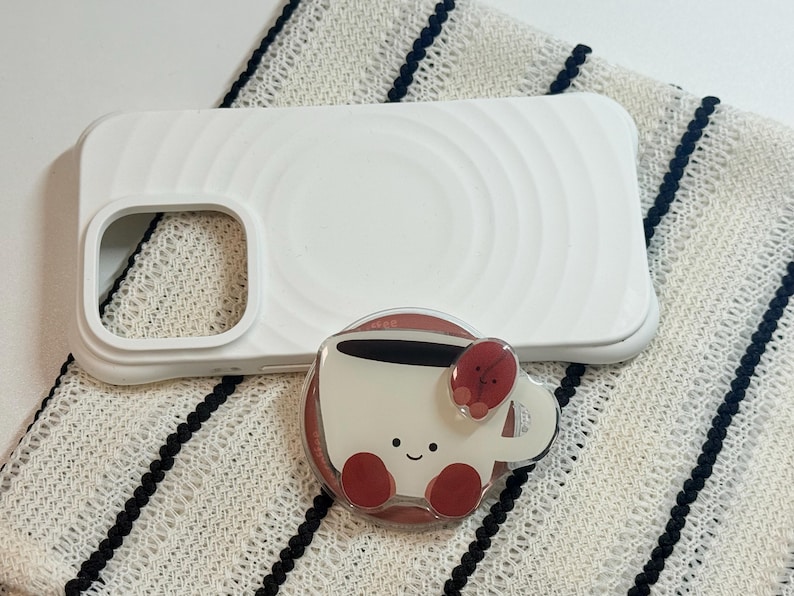 Może przedstawiać: Białe etui na telefon z okrągłym wzorem i uchwytem na telefon w kształcie filiżanki kawy. Uchwyt na telefon jest biały z uśmiechniętą twarzą, czerwonymi akcentami i wzorem ziarna kawy.