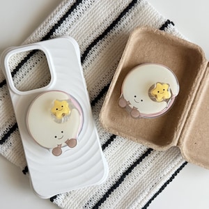 May include: A white smartphone case with a wavy texture and a circular phone grip featuring a smiling white moon character with a small yellow star. A matching circular phone grip is placed inside a small, open, light brown cardboard container.