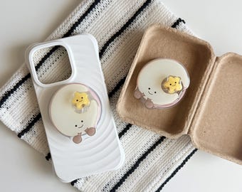 May include: A white smartphone case with a wavy texture and a circular phone grip featuring a smiling white moon character with a small yellow star. A matching circular phone grip is placed inside a small, open, light brown cardboard container.