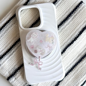 May include: A white smartphone case featuring a heart-shaped attachment with a pearlescent finish. The heart displays illustrations of small animals, a house, and stars, with a dangling pink ribbon charm attached to the bottom edge of the heart.