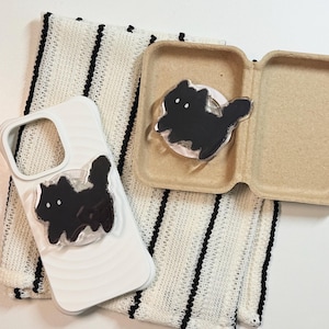 May include: A white phone case with a black cat design, a matching cat phone grip, and a cat-themed phone grip in a cardboard container, all on a white and black striped cloth.