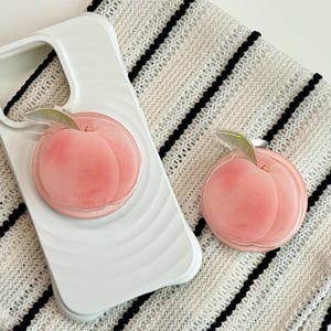 Könnte beinhalten: Weißes Handycase und passender Handyhalter mit einem rosa Pfirsichdesign und einem grünen Blatt. Das Zubehör liegt auf einem weiß-schwarz gestreiften Stoff. Das Handycase hat ein geschwungenes Design.