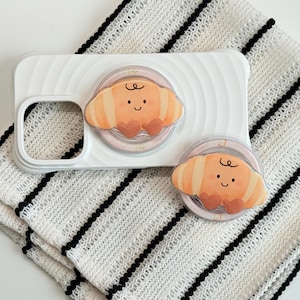 May include: A white phone case with a pop-up grip featuring a cartoon croissant design. Two matching pop-up grips are also visible. The grips have a light orange and pink color scheme with a smiling croissant face. The items are on a white and black striped cloth.