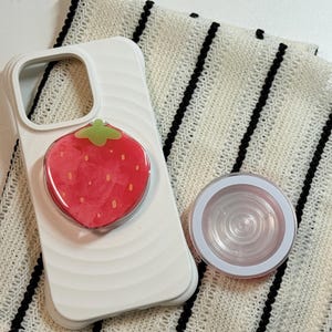 Pu&ograve; includere: Custodia per telefono bianca con un pop-socket a fragola rossa e un'impugnatura per telefono rotonda e trasparente. Il pop-socket a fragola ha un gambo verde. Gli articoli sono su un tessuto a righe bianche e nere.