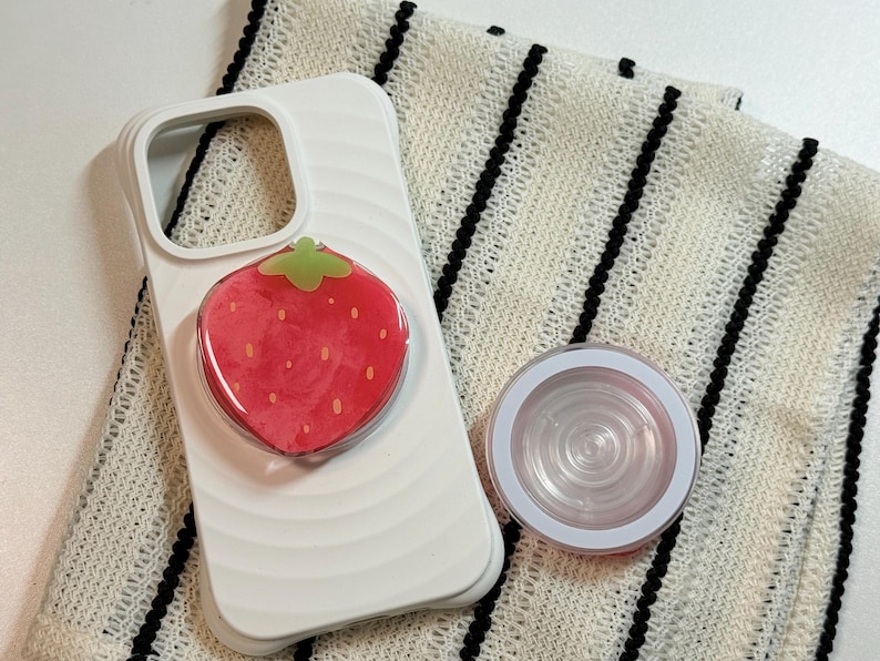 Pu&ograve; includere: Custodia per telefono bianca con un pop-socket a fragola rossa e un'impugnatura per telefono rotonda e trasparente. Il pop-socket a fragola ha un gambo verde. Gli articoli sono su un tessuto a righe bianche e nere.