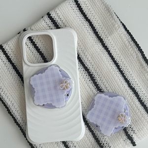 Può includere: Custodia per telefono bianca con un design ondulato, abbinata a un pop socket coordinato. Il pop socket e la custodia presentano un motivo a quadretti lilla chiaro con un accento dorato. Gli articoli sono su un tessuto bianco e nero a righe.