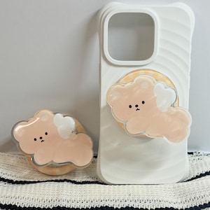 May include: A white phone case with a cute peach-coloured bear design and a matching phone grip. The bear has a white heart-shaped wing and small black dots for eyes. The phone grip is attached to a wooden ring.