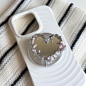May include: A white phone case featuring a circular, silver-toned, heart-shaped mirror attachment. The mirror frame has a metallic, liquid-like drip texture and is accented with three small, light pink, heart-shaped crystals embedded in the metal.