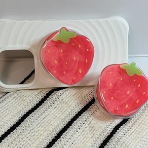 Pu&ograve; includere: Custodia per telefono bianca con design a fragola. La custodia ha un'impugnatura trasparente e lucida a forma di fragola. Le fragole sono rosse con cime verdi e piccoli semi bianchi.