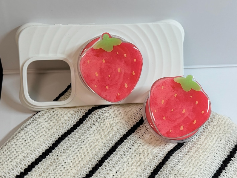 Pu&ograve; includere: Custodia per telefono bianca con design a fragola. La custodia ha un'impugnatura trasparente e lucida a forma di fragola. Le fragole sono rosse con cime verdi e piccoli semi bianchi.