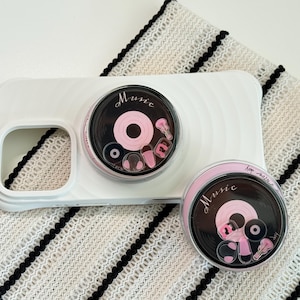 Könnte beinhalten: Weißes Handycase mit einem schwarz-rosa Pop-Socket zum Thema Musik. Der Pop-Socket hat ein Vinyl-Schallplatten-Design mit dem Wort "Music" und anderen Musik-Symbolen. Der Hintergrund ist ein weiß-schwarz gestreifter Stoff.