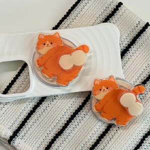 May include: White phone case featuring an orange cartoon cat design, with a matching phone grip. The cat is orange with white details and a friendly expression. The phone case and grip are on a white and black striped cloth.