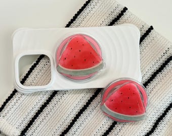 Può includere: Una custodia per telefono bianca con un design a forma di anguria e due pop socket abbinati. Il design dell'anguria presenta una fetta rossa con semi neri e una buccia verde. I pop socket sono rotondi e trasparenti.