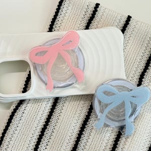Könnte beinhalten: Eine weiße Handyhülle mit einem rosa schleifenförmigen Handygriff, zusammen mit einem separaten blauen schleifenförmigen Griff. Die Griffe haben ein glitzerndes Finish. Die Artikel liegen auf einem weiß-schwarz gestreiften Stoff.