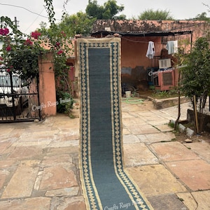 Peut inclure: Un long tapis rectangulaire avec un centre bleu-vert et une bordure beige ornée d'un motif de losanges. Le tapis est exposé à l'extérieur sur un patio en pierre, mettant en valeur sa longueur et son design. Les dimensions du tapis ne sont pas visibles.