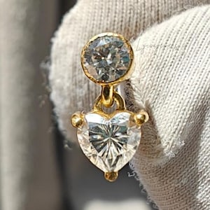 Könnte beinhalten: Ein goldener Ohrring mit zwei klaren, funkelnden Edelsteinen. Der obere Stein ist rund, der untere herzförmig. Der Ohrring wird vor einem weißen Stoffhintergrund gehalten.