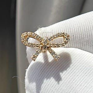 May include: A gold-coloured bow-shaped stud earring, adorned with small, clear gemstones. The earring is set against a white fabric background, with shadows cast by the light. The bow design is intricate, highlighting the sparkle of the stones.