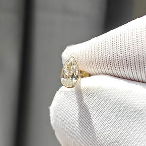 Op de afbeelding: Een close-up van een peervormige diamanten ring. De diamant heeft een lichtgele tint en is gezet in een gouden band. De ring wordt vastgehouden door een gehandschoende hand tegen een neutrale achtergrond.