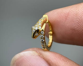 May include: A gold hoop earring shaped like a snake, featuring a textured head and body adorned with small, clear, sparkling stones. The snake design curves to form the circular hoop, with the head resting against the side of the hoop band.