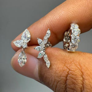May include: Three silver-toned earrings featuring clear, faceted stones. One earring has a butterfly-like shape with a dangling marquise stone, one is a curved climber with marquise stones, and one is a small hoop set with round stones.