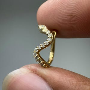 Op de afbeelding: Een gouden oorring in de vorm van een kronkelende slang, voorzien van een reeks kleine, heldere, rondgeslepen stenen langs de bovenste curve van het lichaam. Het metaal heeft een gepolijste afwerking en een S-vormig silhouet.