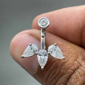 Puede incluir: Un pendiente tipo ear jacket plateado con un perno circular de piedra transparente y una pieza desmontable con tres piedras transparentes en forma de pera dispuestas en abanico, unidas por una barra vertical con dos orificios.
