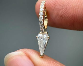 May include: A gold hoop earring featuring a row of small round clear stones along the band and a single, larger, triangular-shaped clear stone dangling from the bottom. The metal is yellow gold, and the stones are set in a polished, reflective finish.