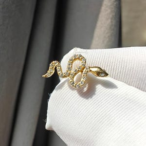 May include: A gold snake ring featuring a coiled body and a small head. The snake's body is adorned with small, clear gemstones. The ring is set against a white fabric background.