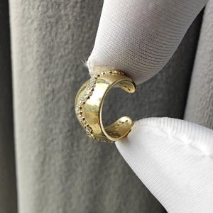 May include: A gold-colored ear cuff with a curved design. The cuff is adorned with small, sparkling gemstones along the outer edge. The jewelry is held by a gloved hand against a gray background.