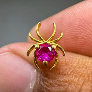 May include: A gold-toned spider-shaped jewelry piece featuring a round, faceted, vibrant pink gemstone at its center. The metal frame forms the spider's body and legs, with the gemstone serving as the abdomen. The design is symmetrical and polished.