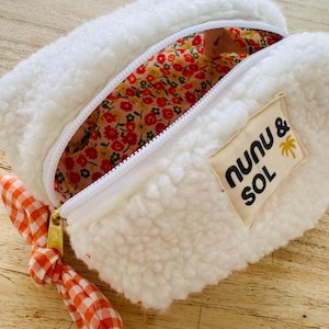 Peut inclure: Trousse blanche et moelleuse avec un intérieur floral et une fermeture éclair blanche. La trousse porte une étiquette "Nunu & Sol" avec un palmier. La tirette de la fermeture éclair est en vichy orange et blanc.