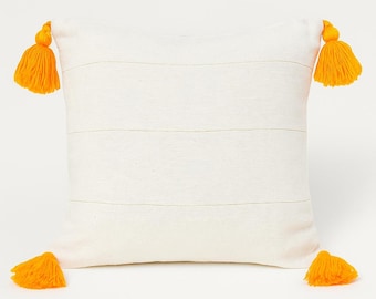 May include: Off-white square pillow with horizontal stripes and four bright yellow tassels. The tassels are located at each corner of the pillow. The pillow is a decorative home accent.
