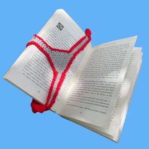 Könnte beinhalten: Ein handgefertigtes Häkel-Lesezeichen mit einem dreieckigen grauen Mittelteil und einem leuchtend roten, gewellten Rand. Das Lesezeichen liegt auf den offenen Seiten eines Buches, wobei der Text auf den weißen Papierseiten unter dem Garn deutlich sichtbar ist.
