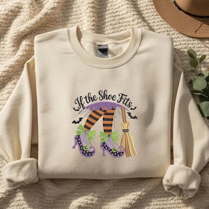 May include: A cream-colored crewneck sweatshirt featuring a graphic print of witch legs in orange and black striped stockings, purple shoes with green bows, and a broomstick. The text above the graphic reads, If the Shoe Fits, in black.