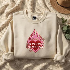 Puede incluir: Una sudadera de cuello redondo color crema con un diseño bordado de un corazón rojo rodeado de llamas rosas. El texto dice: warning: may contain EXPLICIT CONTENT, acompañado de pequeños iconos de estrellas en el centro del pecho.